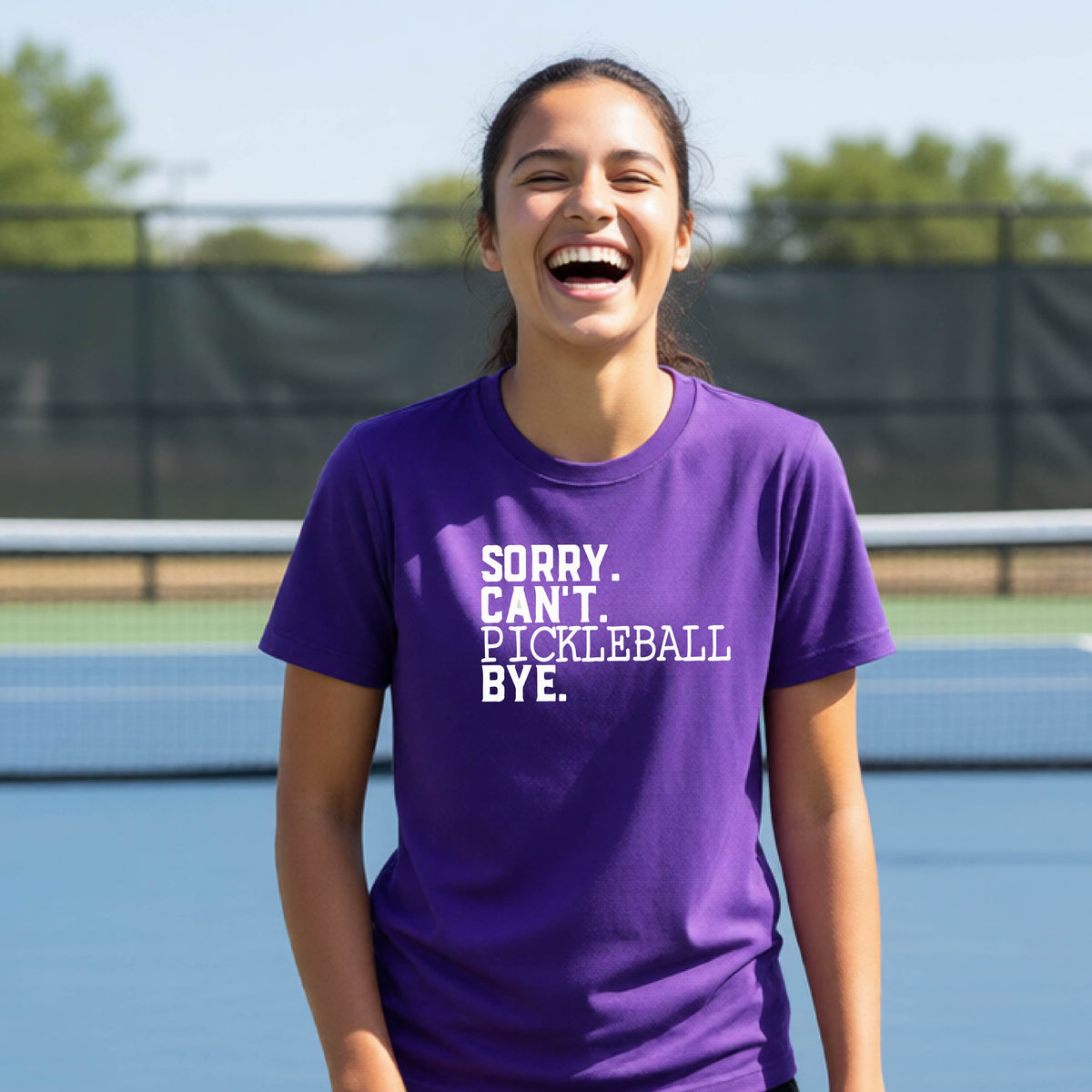A young person on a pickleball court wearing a purple short-sleeve t-shirt with a white graphic of the words  “SORRY. CAN’T. PICKLEBALL BYE” each on a separate line, on the front of the shirt. 