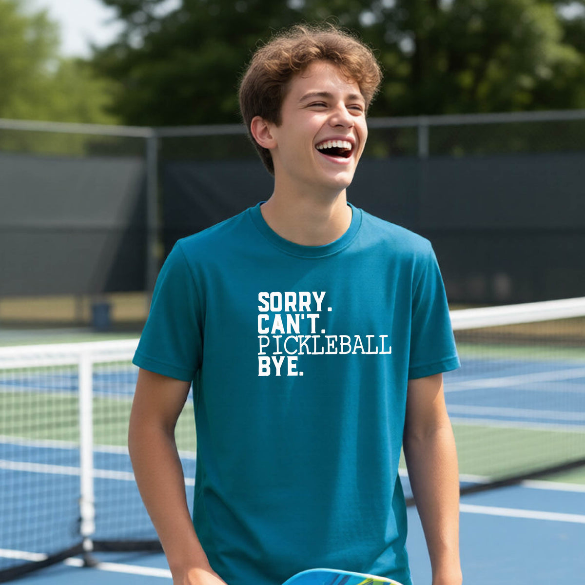 A young person on a pickleball court wearing a teal short-sleeve t-shirt with a white graphic of the words  “SORRY. CAN’T. PICKLEBALL BYE” each on a separate line, on the front of the shirt. 