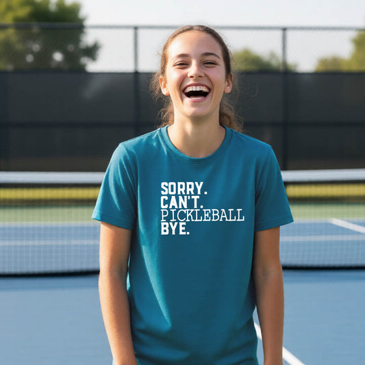 A young person on a pickleball court wearing a teal short-sleeve t-shirt with a white graphic of the words  “SORRY. CAN’T. PICKLEBALL BYE” each on a separate line, on the front of the shirt. 