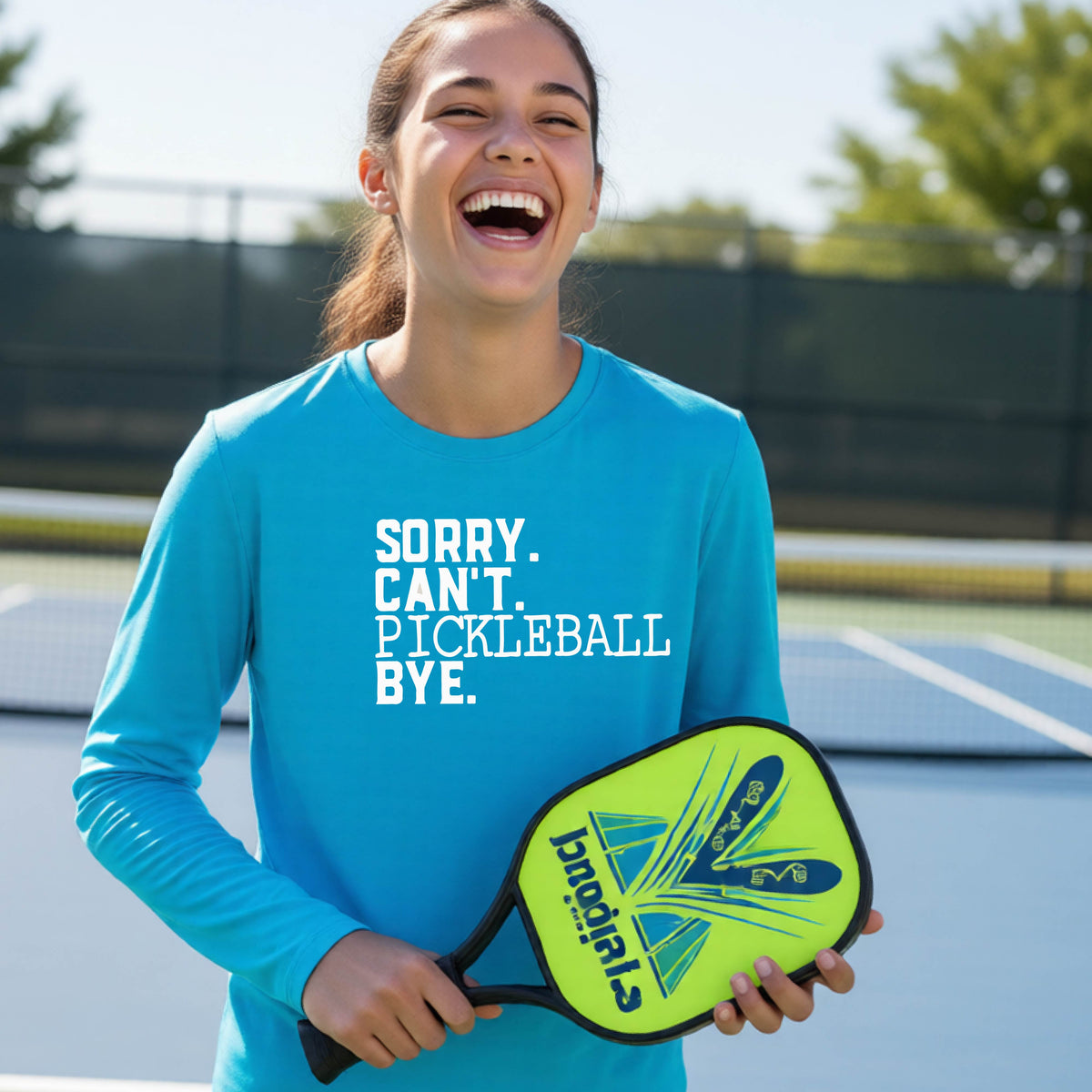 A young person on a pickleball court wearing a bright blue short-sleeve t-shirt with a white graphic of the words  “SORRY. CAN’T. PICKLEBALL BYE” each on a separate line, on the front of the shirt.