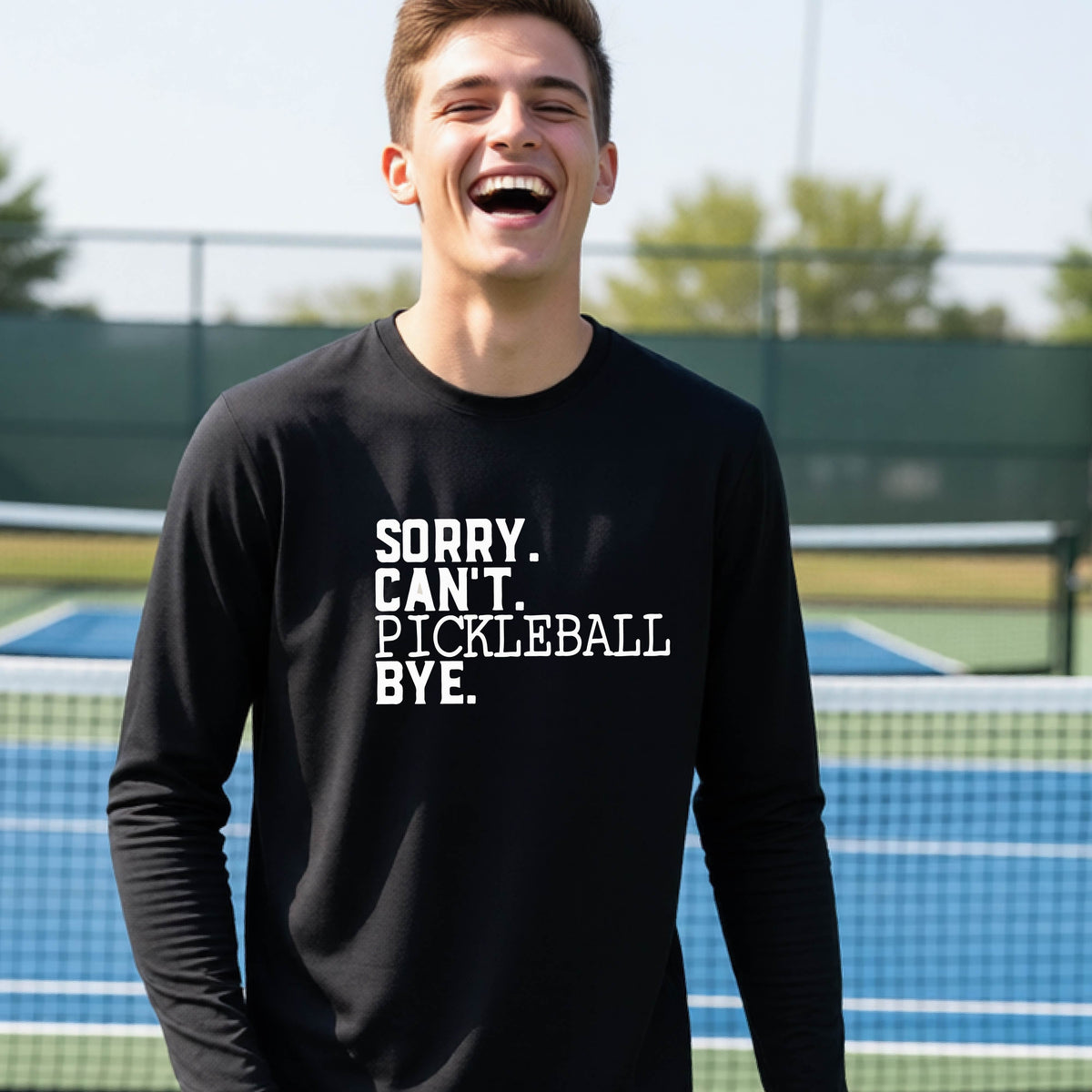 A young person on a pickleball court wearing a black short-sleeve t-shirt with a white graphic of the words  “SORRY. CAN’T. PICKLEBALL BYE” each on a separate line, on the front of the shirt. 