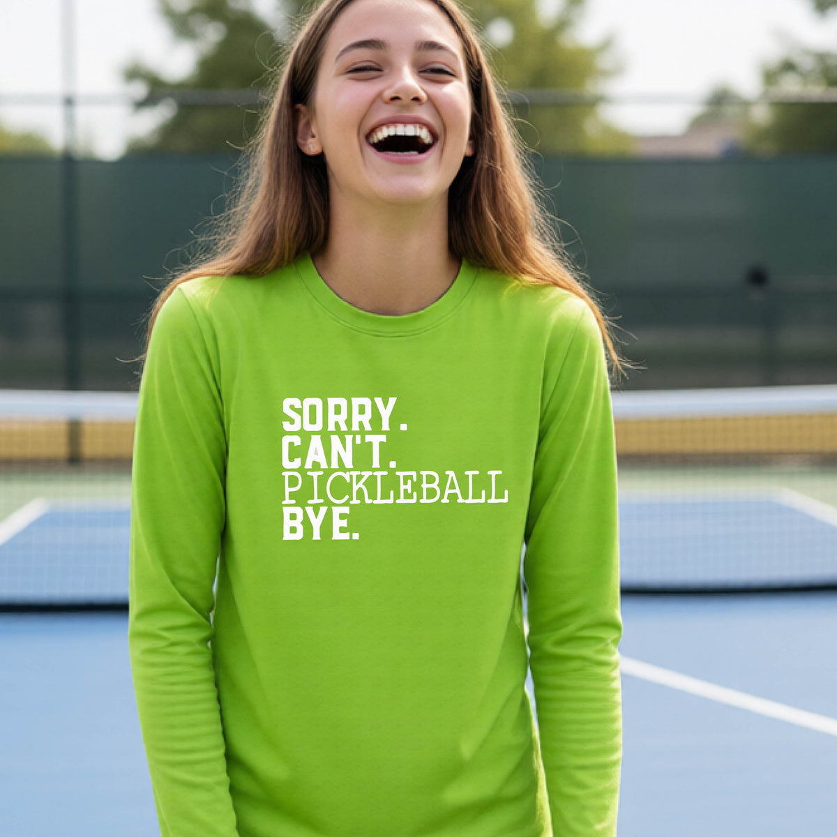 A young person on a pickleball court wearing a lime green short-sleeve t-shirt with a white graphic of the words  “SORRY. CAN’T. PICKLEBALL BYE” each on a separate line, on the front of the shirt.