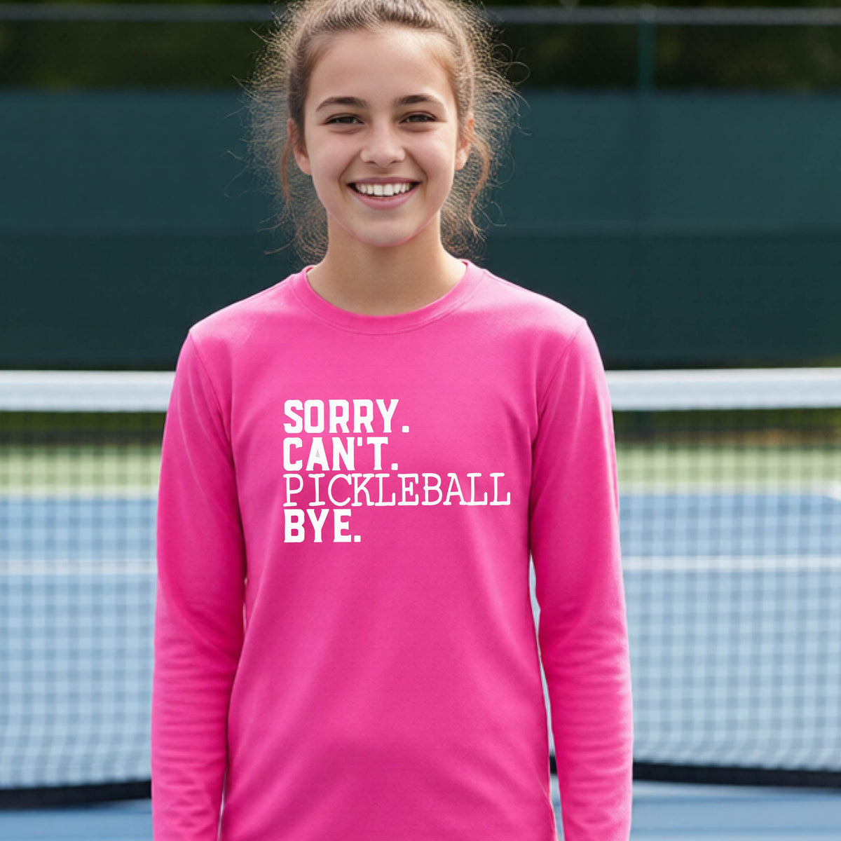 A young person on a pickleball court wearing a pink short-sleeve t-shirt with a white graphic of the words  “SORRY. CAN’T. PICKLEBALL BYE” each on a separate line, on the front of the shirt