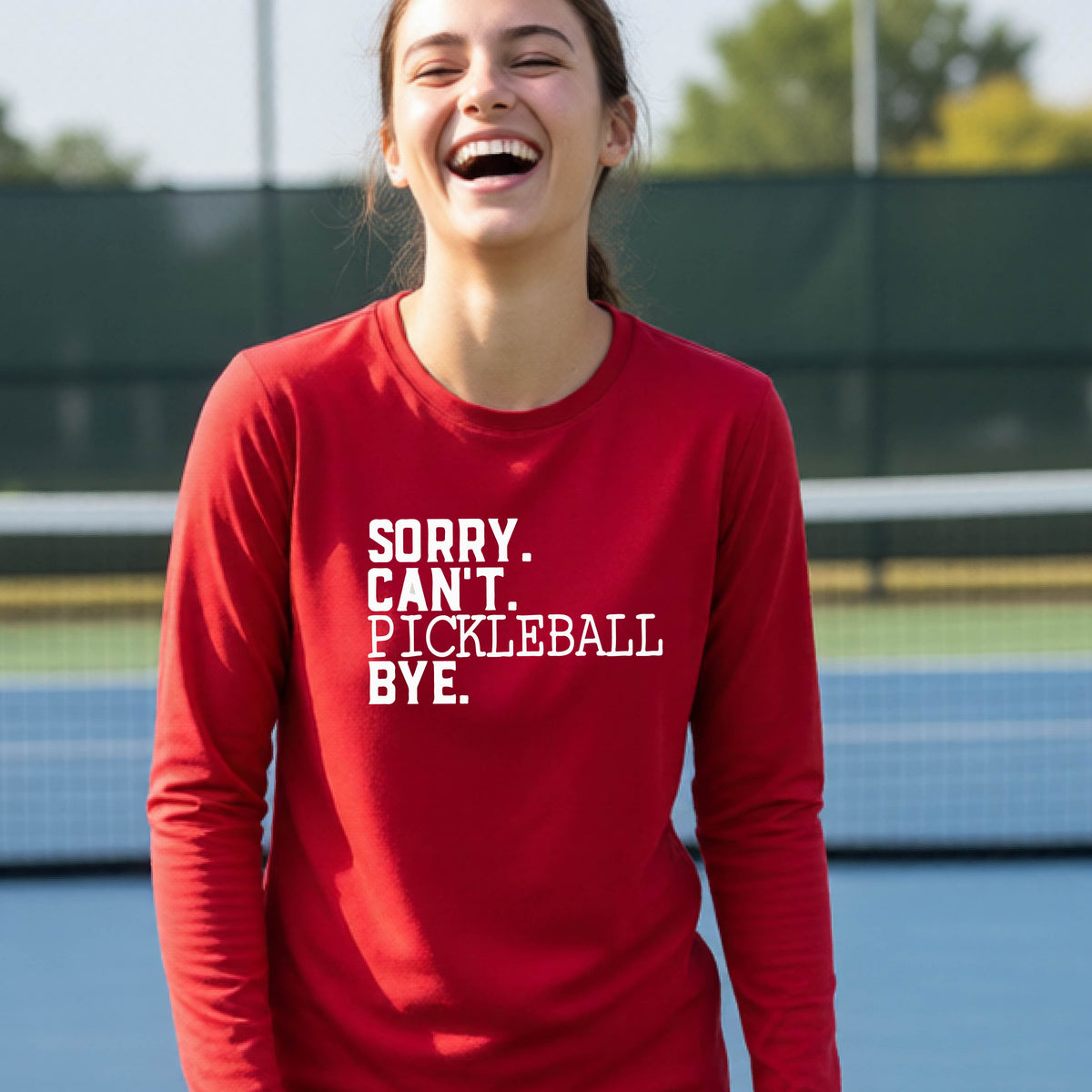 A young person on a pickleball court wearing a red short-sleeve t-shirt with a white graphic of the words  “SORRY. CAN’T. PICKLEBALL BYE” each on a separate line, on the front of the shirt.