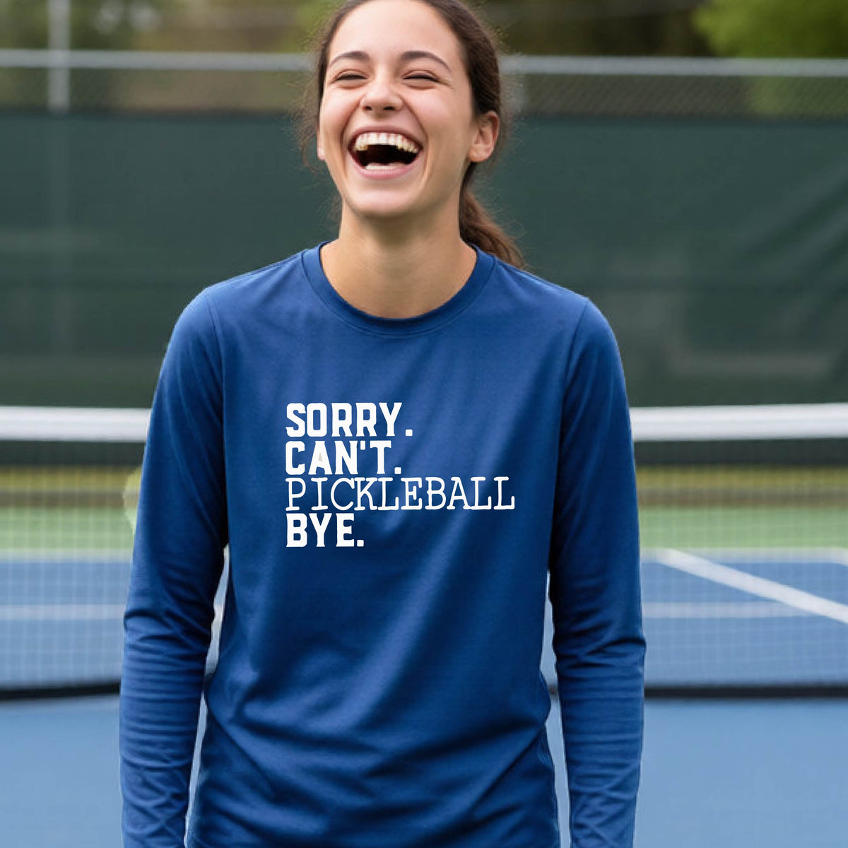 A young person on a pickleball court wearing a royal blue short-sleeve t-shirt with a white graphic of the words  “SORRY. CAN’T. PICKLEBALL BYE” each on a separate line, on the front of the shirt.
