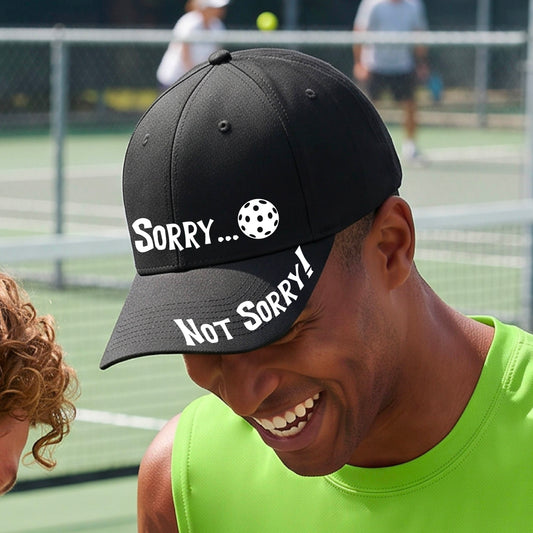 A close-up of a black mid-profile athletic hat with pickleball motif design on the front and brim.  The front features the playful word “SORRY…” in bold white lettering, accented a colorful pickleball, and the brim design features “NOT SORRY!” in bold white lettering curved around the brim edge.