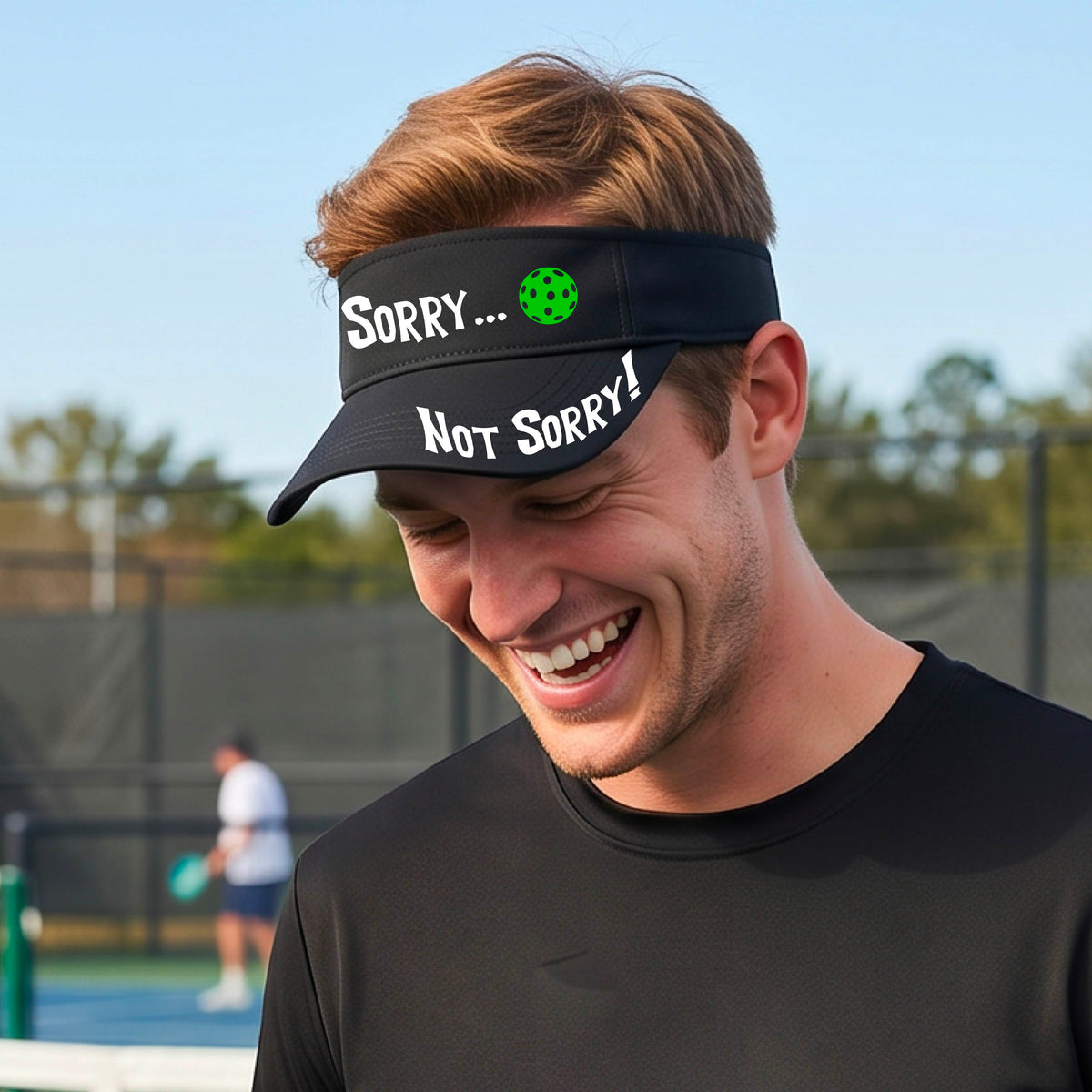 A close-up of a black athletic visor with motif.design on the front and brim..  The front features the playful word “SORRY…” in bold white lettering, accented a colorful pickleball, and the brim design features “NOT SORRY!” in bold white lettering curved around the brim edge.