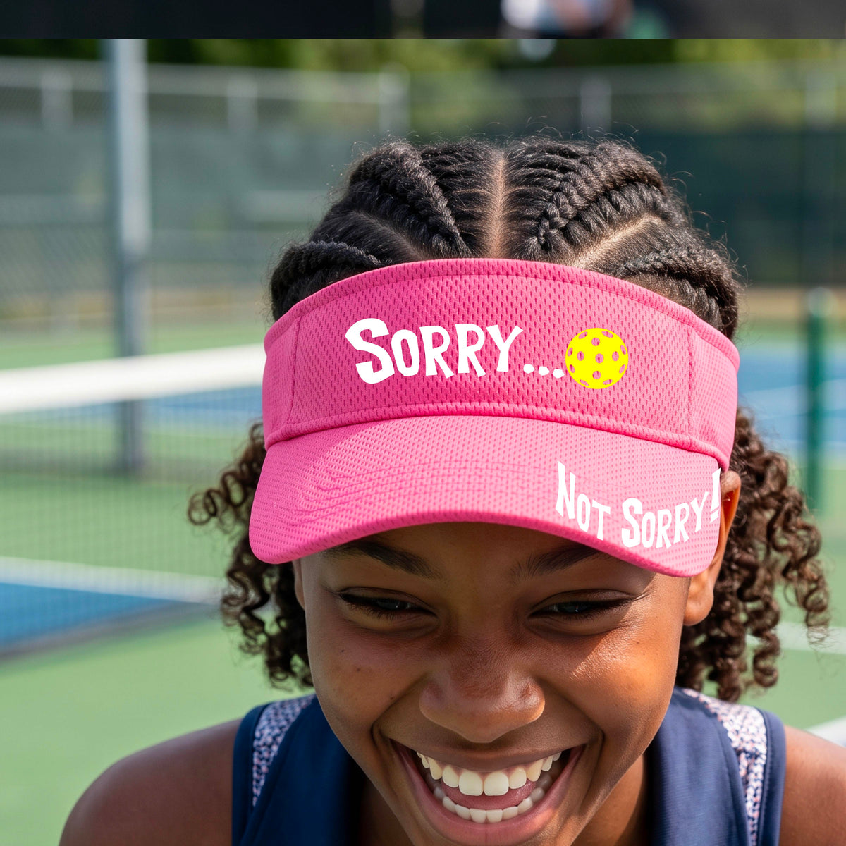 A close-up of a pink athletic visor with motif.design on the front and brim..  The front features the playful word “SORRY…” in bold white lettering, accented a colorful pickleball, and the brim design features “NOT SORRY!” in bold white lettering curved around the brim edge.
