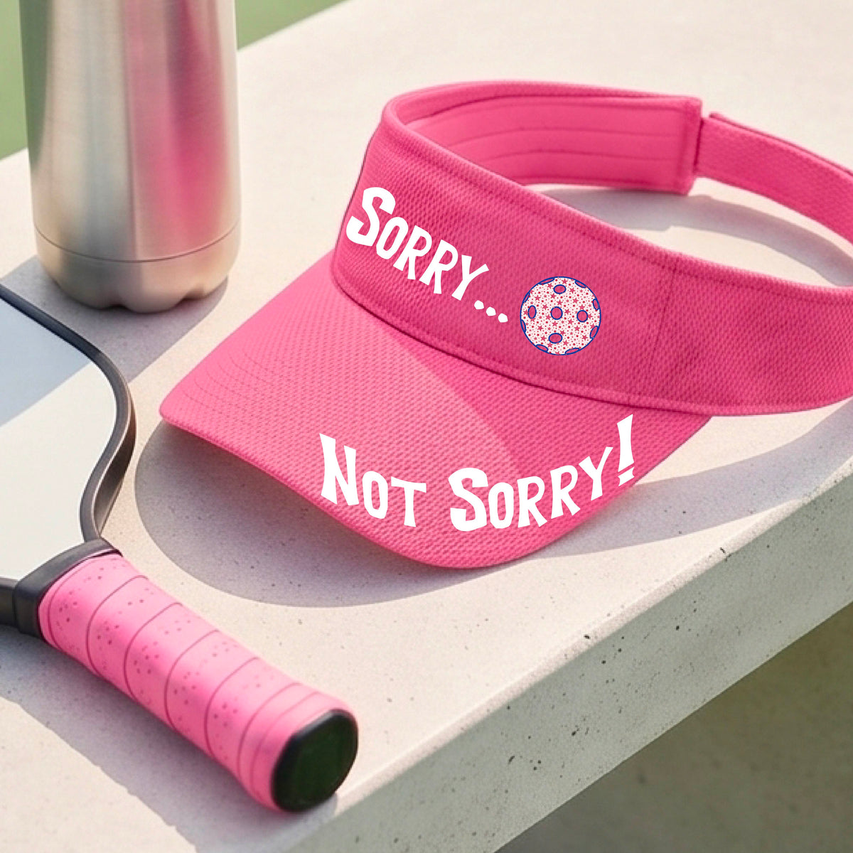 A close-up of a pink athletic visor with motif.design on the front and brim..  The front features the playful word “SORRY…” in bold white lettering, accented a colorful pickleball, and the brim design features “NOT SORRY!” in bold white lettering curved around the brim edge.