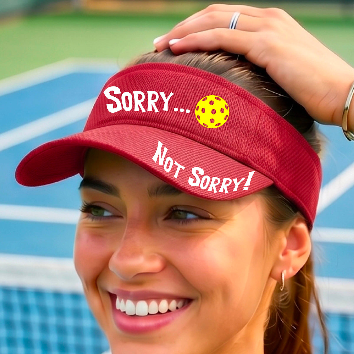 A close-up of a red athletic visor with motif.design on the front and brim..  The front features the playful word “SORRY…” in bold white lettering, accented a colorful pickleball, and the brim design features “NOT SORRY!” in bold white lettering curved around the brim edge.