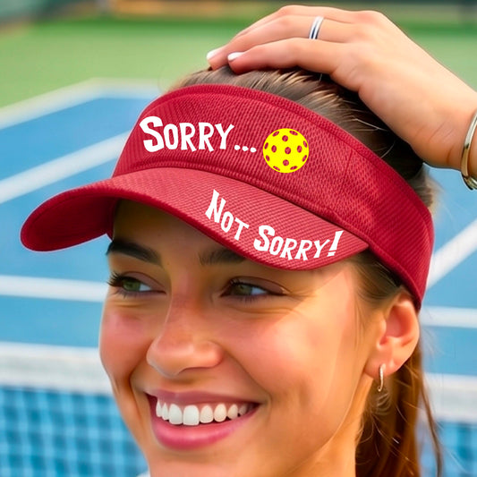 A close-up of a red athletic visor with motif.design on the front and brim..  The front features the playful word “SORRY…” in bold white lettering, accented a colorful pickleball, and the brim design features “NOT SORRY!” in bold white lettering curved around the brim edge.