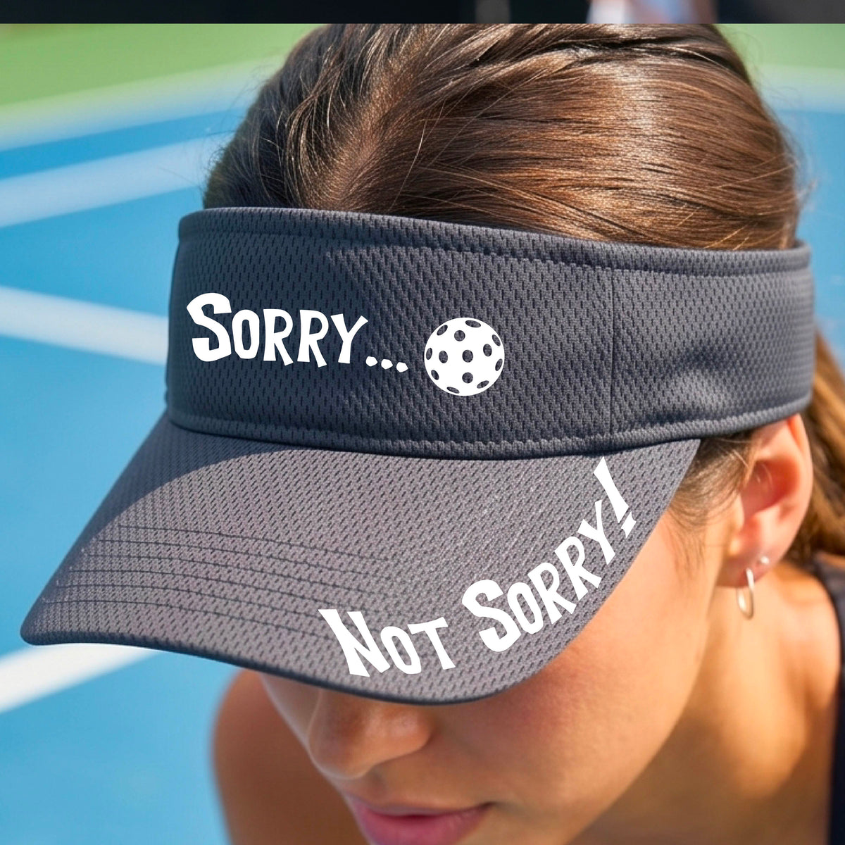 A close-up of a graphite gray athletic visor with motif.design on the front and brim..  The front features the playful word “SORRY…” in bold white lettering, accented a colorful pickleball, and the brim design features “NOT SORRY!” in bold white lettering curved around the brim edge.