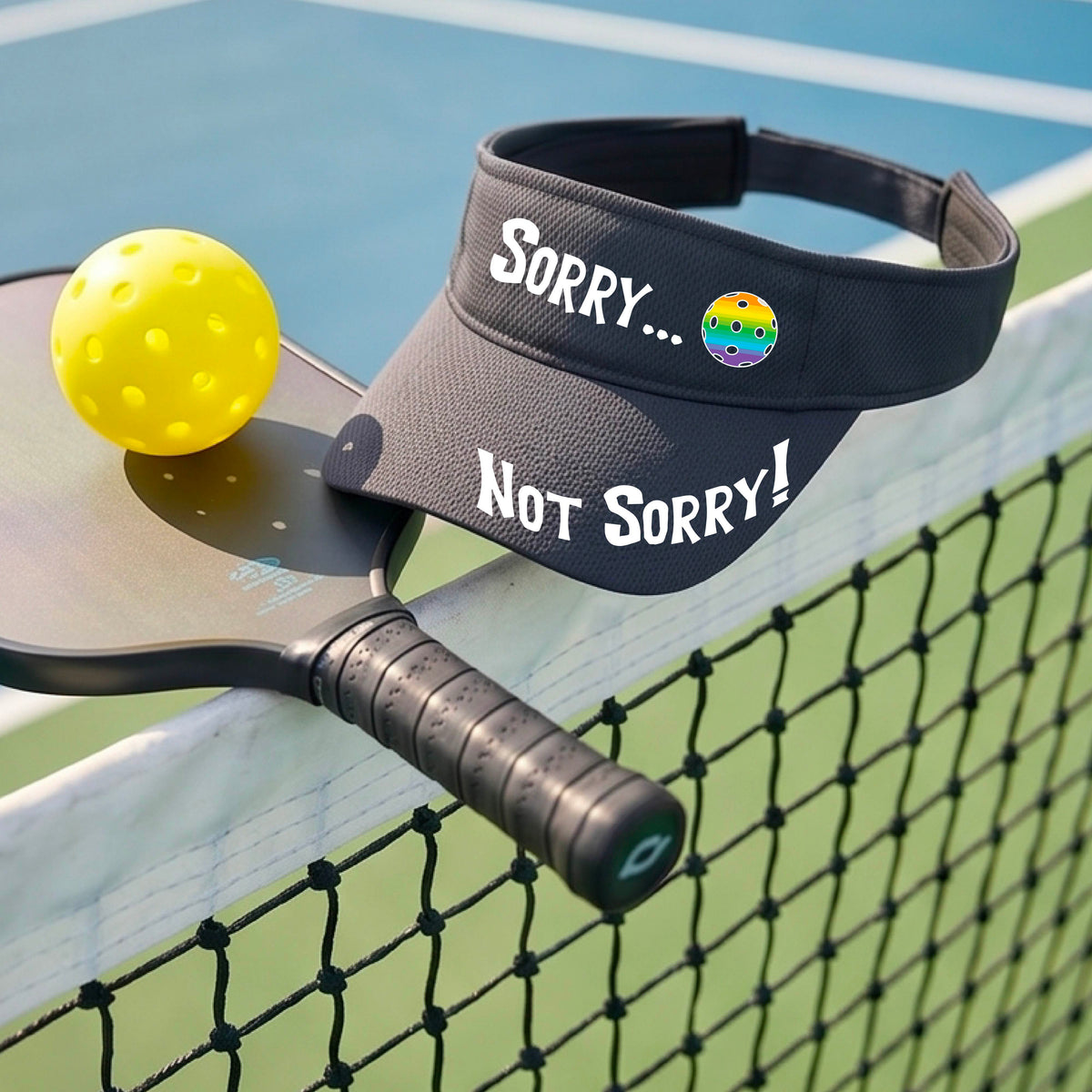 A close-up of a graphite gray athletic visor with motif.design on the front and brim..  The front features the playful word “SORRY…” in bold white lettering, accented a colorful pickleball, and the brim design features “NOT SORRY!” in bold white lettering curved around the brim edge.
