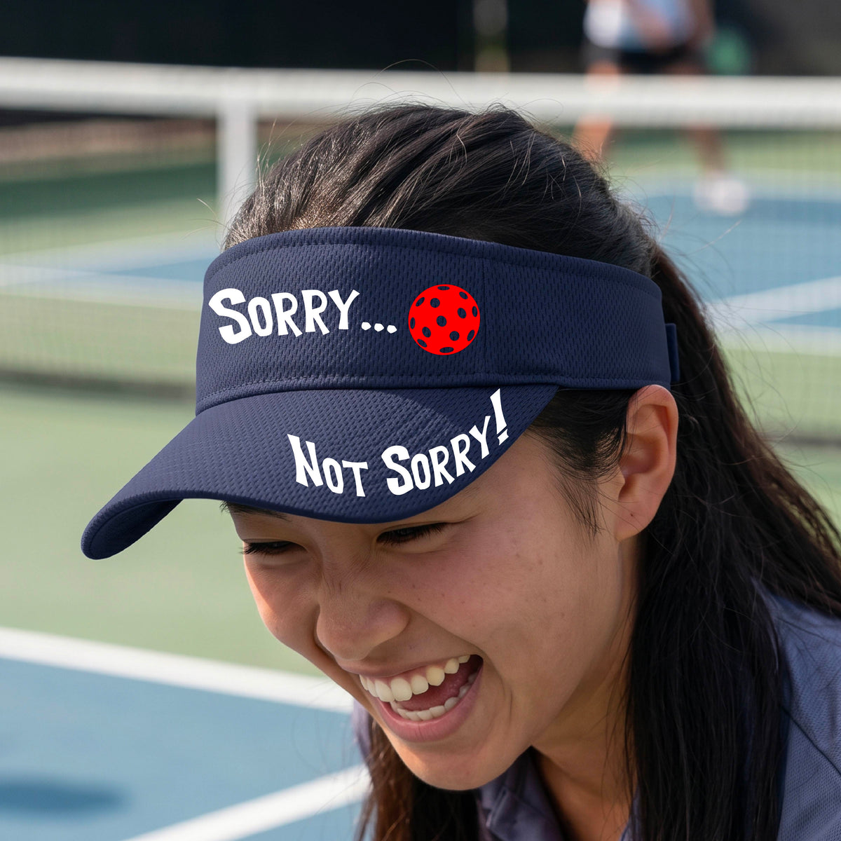 A close-up of a navy blue athletic visor with motif.design on the front and brim..  The front features the playful word “SORRY…” in bold white lettering, accented a colorful pickleball, and the brim design features “NOT SORRY!” in bold white lettering curved around the brim edge.
