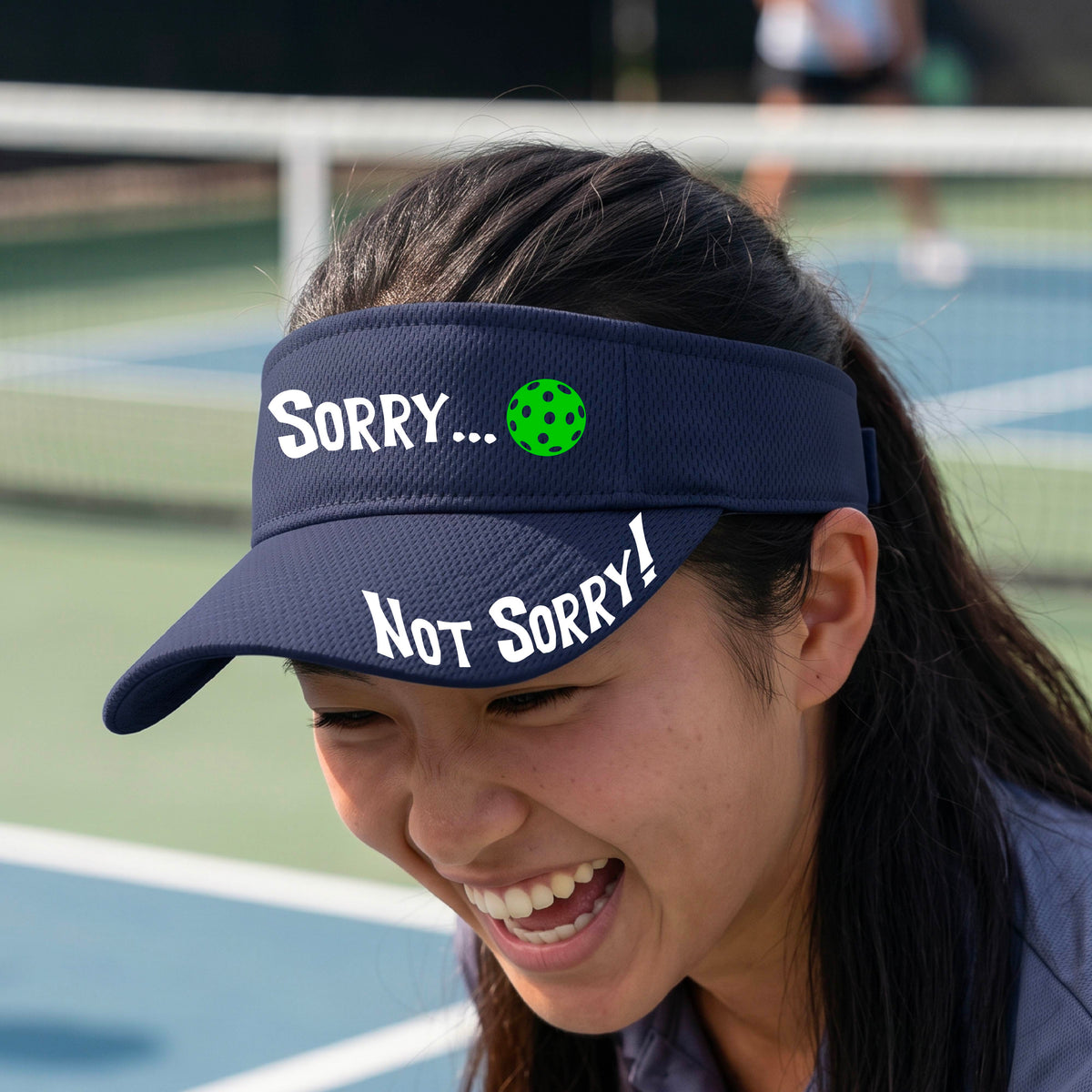 A close-up of a navy blue athletic visor with motif.design on the front and brim..  The front features the playful word “SORRY…” in bold white lettering, accented a colorful pickleball, and the brim design features “NOT SORRY!” in bold white lettering curved around the brim edge.