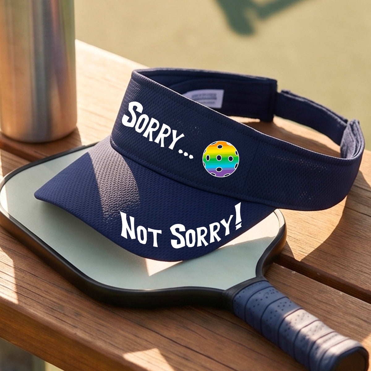 A close-up of a navy blue athletic visor with motif.design on the front and brim..  The front features the playful word “SORRY…” in bold white lettering, accented a colorful pickleball, and the brim design features “NOT SORRY!” in bold white lettering curved around the brim edge.