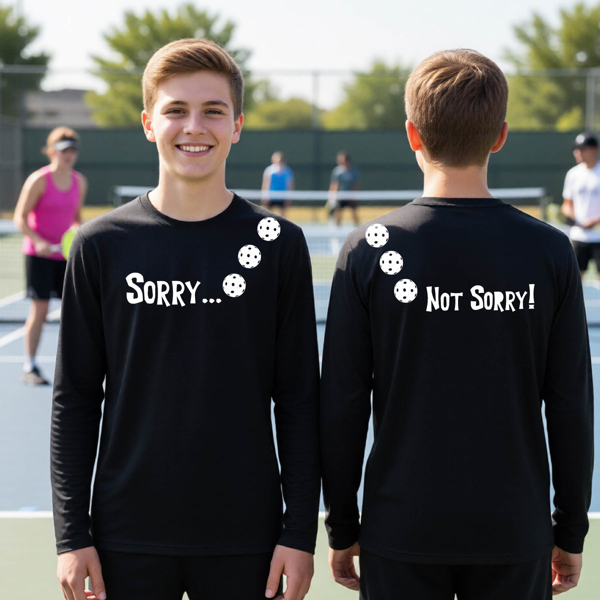 Two young people on a pickleball court wearing black long-sleeve t-shirts with a front/back graphic design.  The front features the playful word “SORRY…” in bold white lettering, accented with three colorful pickleballs arranged diagonally across the upper chest.  The back design features “NOT SORRY!” in bold white lettering running vertically down the upper back, with three matching colorful pickleballs positioned above lettering.  