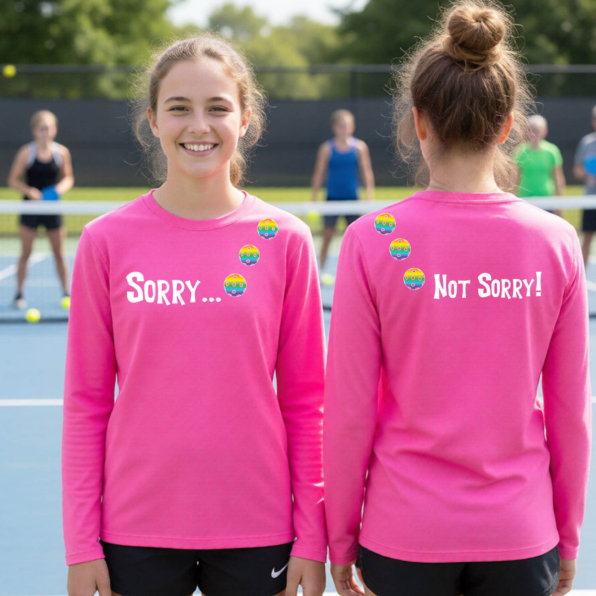 Two young people on a pickleball court wearing pink long-sleeve t-shirts with a front/back graphic design.  The front features the playful word “SORRY…” in bold white lettering, accented with three colorful pickleballs arranged diagonally across the upper chest.  The back design features “NOT SORRY!” in bold white lettering running vertically down the upper back, with three matching colorful pickleballs positioned above lettering.  