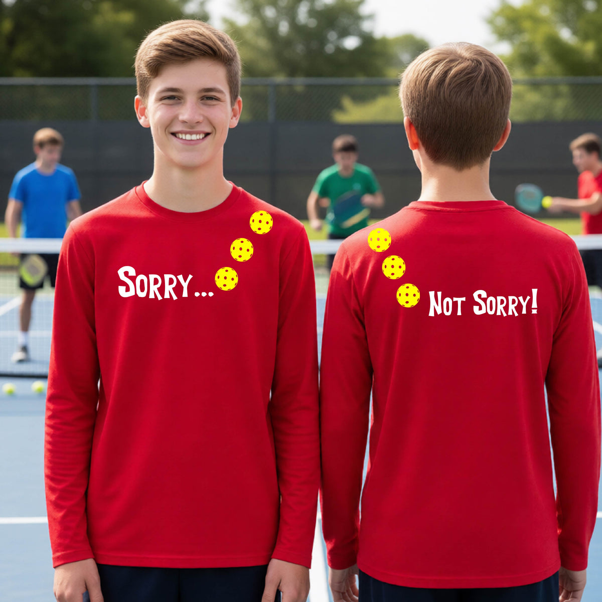 Two young people on a pickleball court wearing red long-sleeve t-shirts with a front/back graphic design.  The front features the playful word “SORRY…” in bold white lettering, accented with three colorful pickleballs arranged diagonally across the upper chest.  The back design features “NOT SORRY!” in bold white lettering running vertically down the upper back, with three matching colorful pickleballs positioned above lettering.  