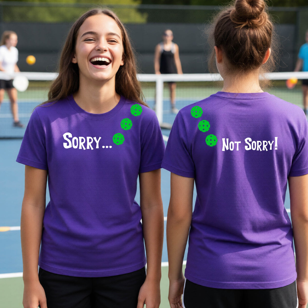 Two youths on a pickleball court wearing purple short-sleeve t-shirts with a front/back graphic design.  The front features the playful word “SORRY…” in bold white lettering, accented with three colorful pickleballs arranged diagonally across the upper chest.  The back design features “NOT SORRY!” in bold white lettering running vertically down the upper back, with three matching colorful pickleballs positioned above lettering.  
