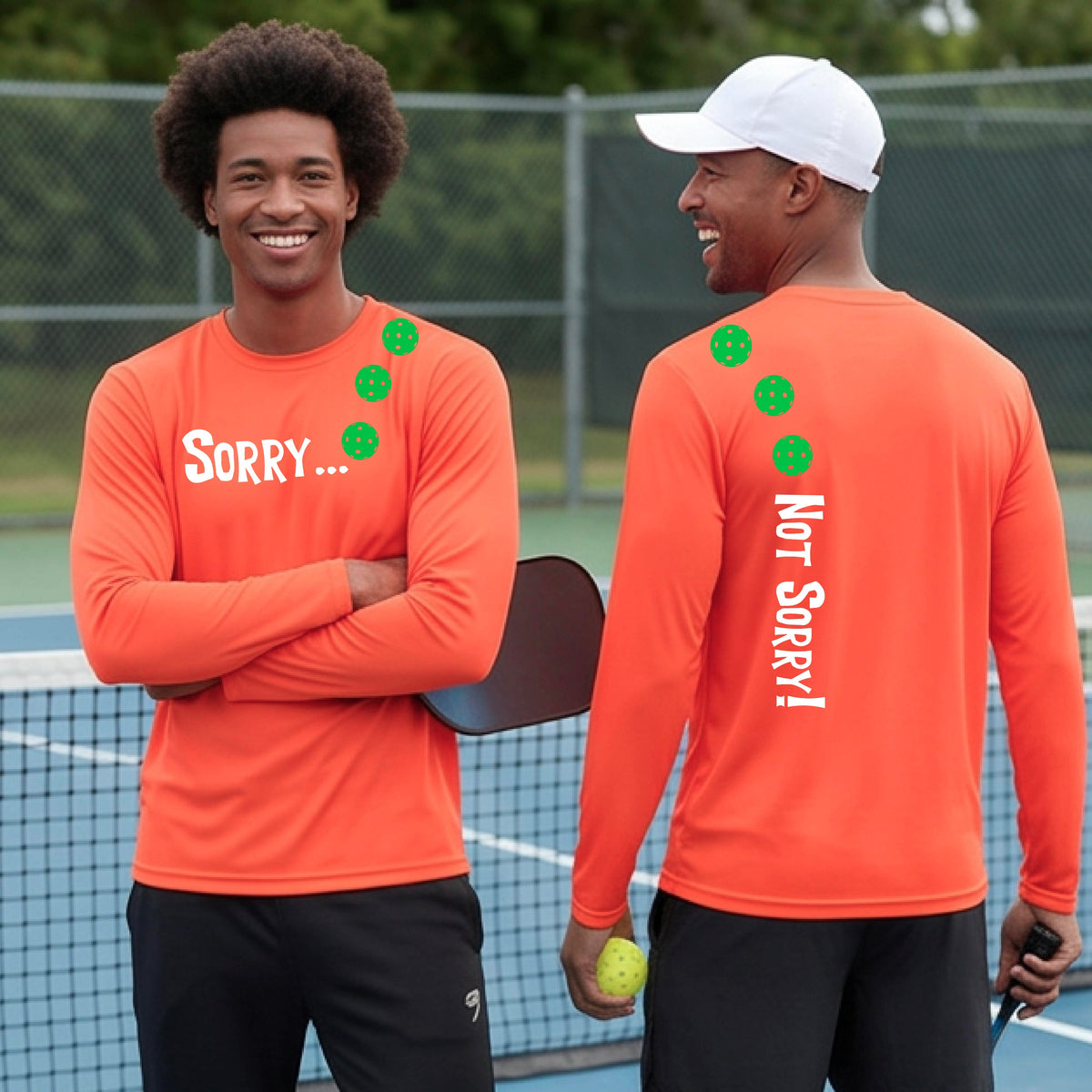Two men on a pickleball court wearing orange long-sleeve t-shirts with a front/back graphic design.  The front features the playful word “SORRY…” in bold white lettering, accented with three colorful pickleballs arranged diagonally across the upper chest.  The back design features “NOT SORRY!” in bold white lettering running vertically down the upper back, with three matching colorful pickleballs positioned above lettering.  