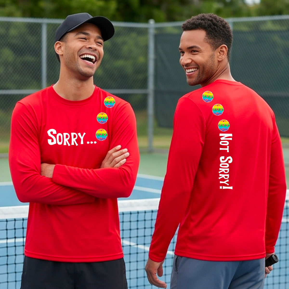 Two men on a pickleball court wearing red long-sleeve t-shirts with a front/back graphic design.  The front features the playful word “SORRY…” in bold white lettering, accented with three colorful pickleballs arranged diagonally across the upper chest.  The back design features “NOT SORRY!” in bold white lettering running vertically down the upper back, with three matching colorful pickleballs positioned above lettering.  
