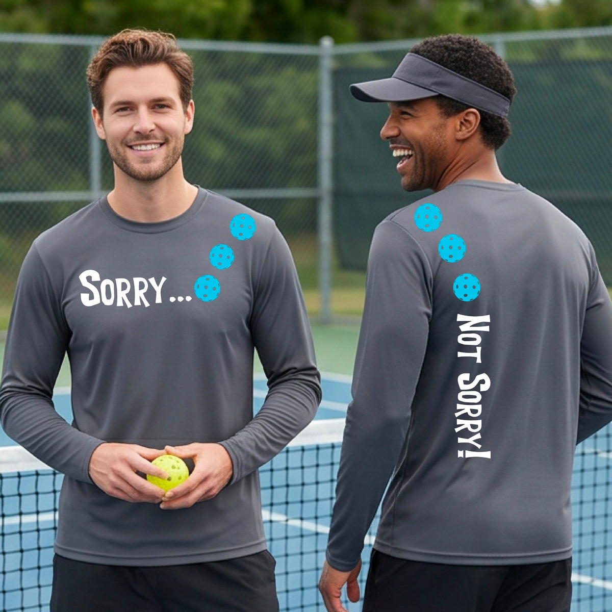 Two men on a pickleball court wearing dark gray long-sleeve t-shirts with a front/back graphic design.  The front features the playful word “SORRY…” in bold white lettering, accented with three colorful pickleballs arranged diagonally across the upper chest.  The back design features “NOT SORRY!” in bold white lettering running vertically down the upper back, with three matching colorful pickleballs positioned above lettering.  