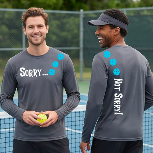 Two men on a pickleball court wearing dark gray long-sleeve t-shirts with a front/back graphic design.  The front features the playful word “SORRY…” in bold white lettering, accented with three colorful pickleballs arranged diagonally across the upper chest.  The back design features “NOT SORRY!” in bold white lettering running vertically down the upper back, with three matching colorful pickleballs positioned above lettering.  
