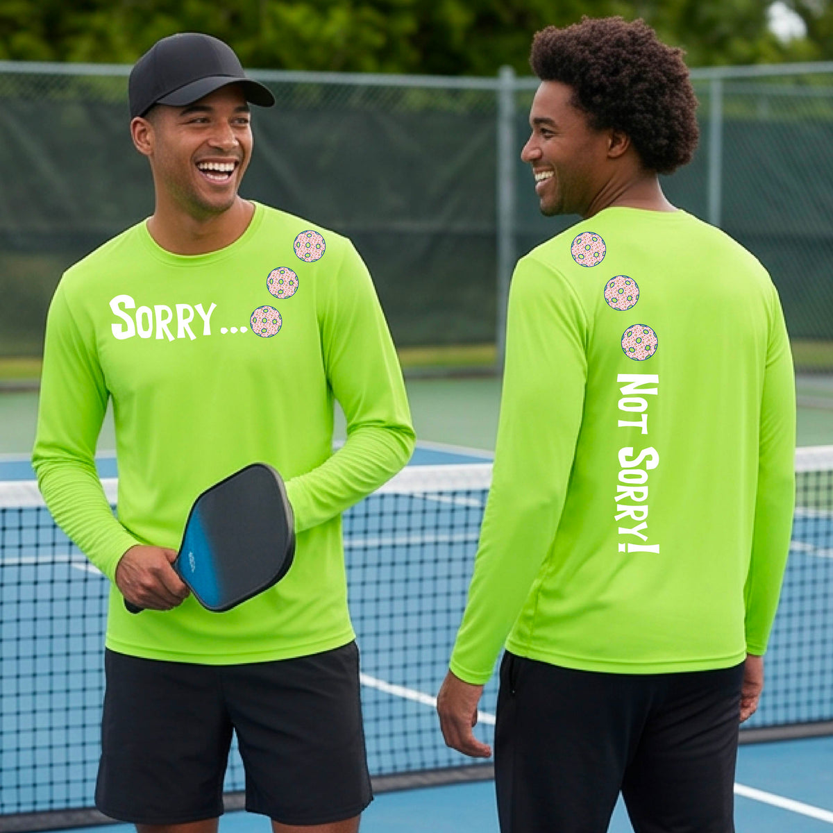 Two men on a pickleball court wearing lime green long-sleeve t-shirts with a front/back graphic design.  The front features the playful word “SORRY…” in bold white lettering, accented with three colorful pickleballs arranged diagonally across the upper chest.  The back design features “NOT SORRY!” in bold white lettering running vertically down the upper back, with three matching colorful pickleballs positioned above lettering.  