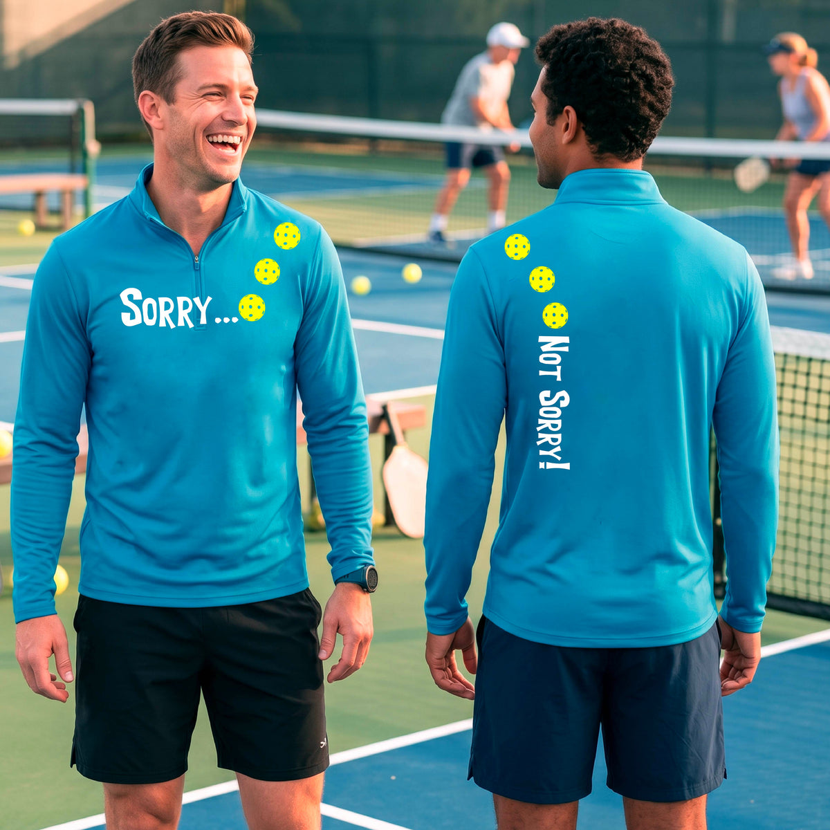 Two men on a pickleball court wearing bright blue quarter-zip shirts with a front/back graphic design.  The front features the playful word “SORRY…” in bold white lettering, accented with three colorful pickleballs arranged diagonally across the upper chest.  The back design features “NOT SORRY!” in bold white lettering running vertically down the upper back, with three matching colorful pickleballs positioned above lettering.  