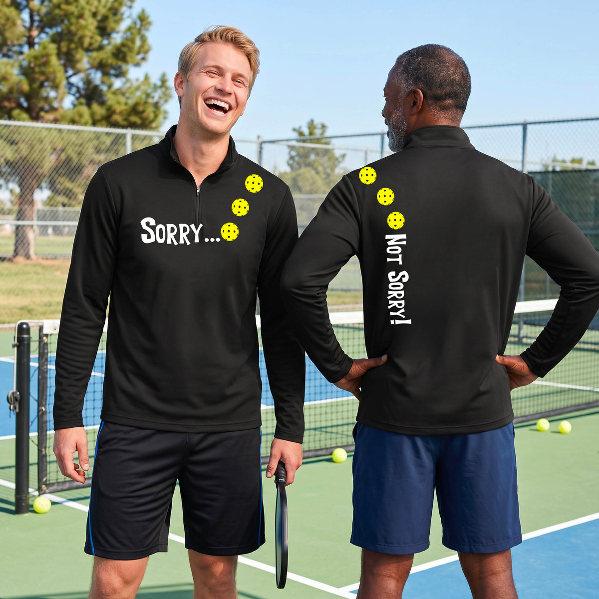 Two men on a pickleball court wearing black quarter-zip shirts with a front/back graphic design.  The front features the playful word “SORRY…” in bold white lettering, accented with three colorful pickleballs arranged diagonally across the upper chest.  The back design features “NOT SORRY!” in bold white lettering running vertically down the upper back, with three matching colorful pickleballs positioned above lettering.  