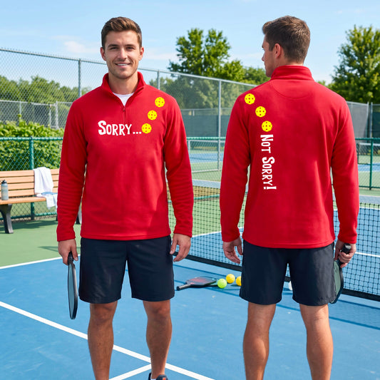 Two men on a pickleball court wearing red quarter-zip shirts with a front/back graphic design.  The front features the playful word “SORRY…” in bold white lettering, accented with three colorful pickleballs arranged diagonally across the upper chest.  The back design features “NOT SORRY!” in bold white lettering running vertically down the upper back, with three matching colorful pickleballs positioned above lettering.  