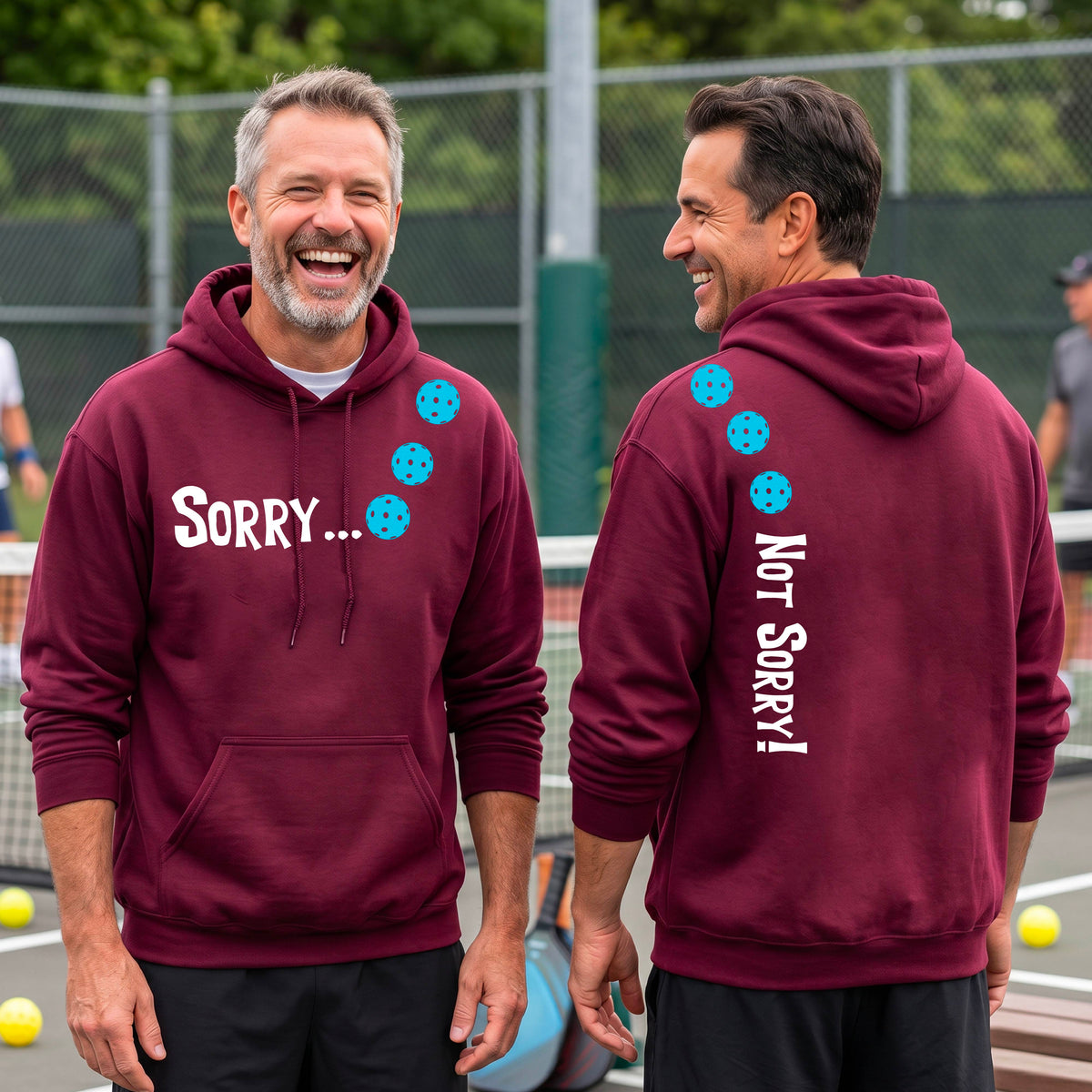 Two men on a pickleball court wearing unisex maroon pullover hoodies with a front/back graphic design.  The front features the playful word “SORRY…” in bold white lettering, accented with three colorful pickleballs arranged diagonally across the upper chest.  The back design features “NOT SORRY!” in bold white lettering running vertically down the upper back, with three matching colorful pickleballs positioned above lettering.  