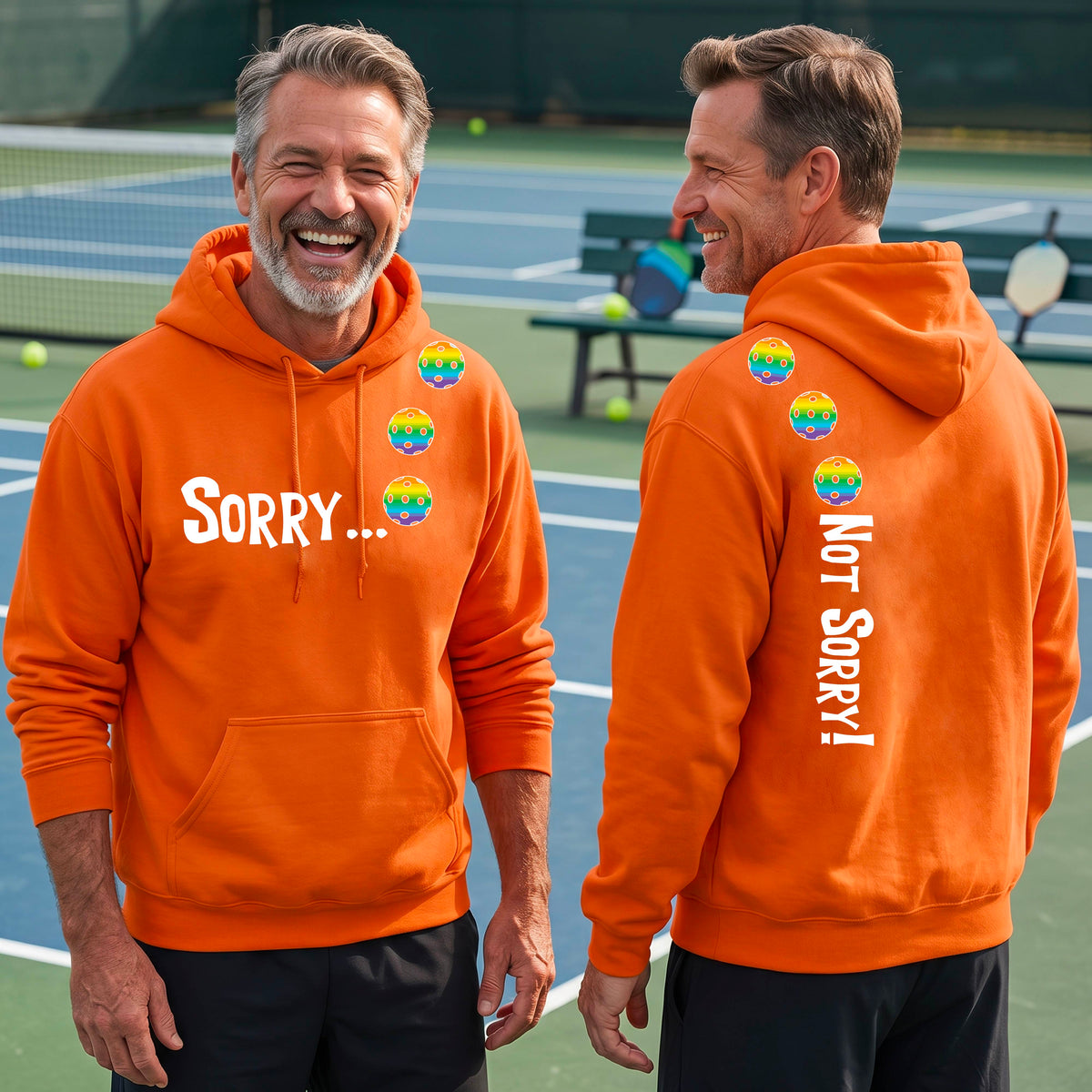 Two men on a pickleball court wearing unisex orange pullover hoodies with a front/back graphic design.  The front features the playful word “SORRY…” in bold white lettering, accented with three colorful pickleballs arranged diagonally across the upper chest.  The back design features “NOT SORRY!” in bold white lettering running vertically down the upper back, with three matching colorful pickleballs positioned above lettering.  