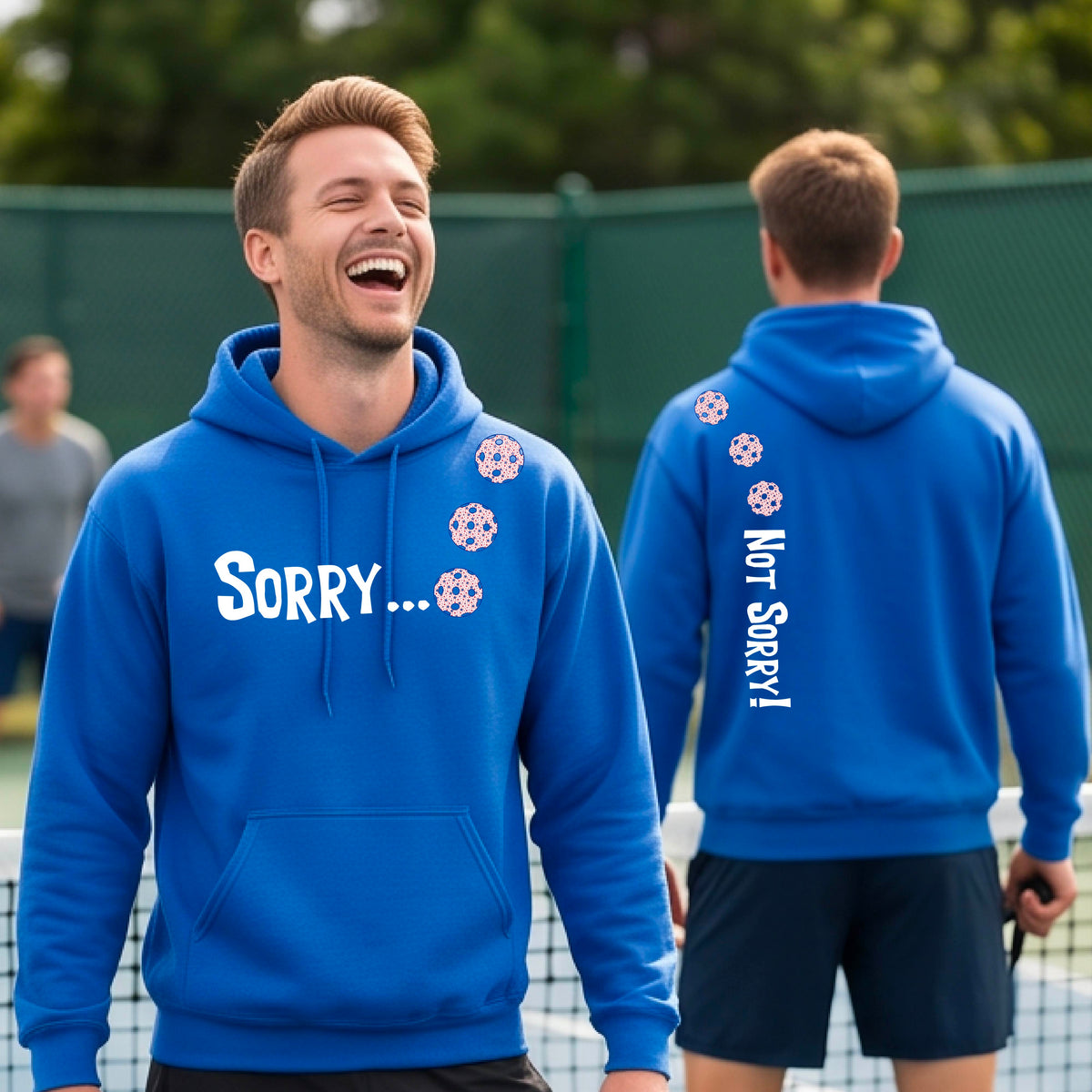 Two men on a pickleball court wearing unisex royal blue pullover hoodies with a front/back graphic design.  The front features the playful word “SORRY…” in bold white lettering, accented with three colorful pickleballs arranged diagonally across the upper chest.  The back design features “NOT SORRY!” in bold white lettering running vertically down the upper back, with three matching colorful pickleballs positioned above lettering.  