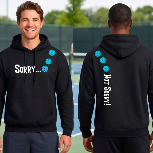 Two men on a pickleball court wearing unisex black pullover hoodies with a front/back graphic design.  The front features the playful word “SORRY…” in bold white lettering, accented with three colorful pickleballs arranged diagonally across the upper chest.  The back design features “NOT SORRY!” in bold white lettering running vertically down the upper back, with three matching colorful pickleballs positioned above lettering.  