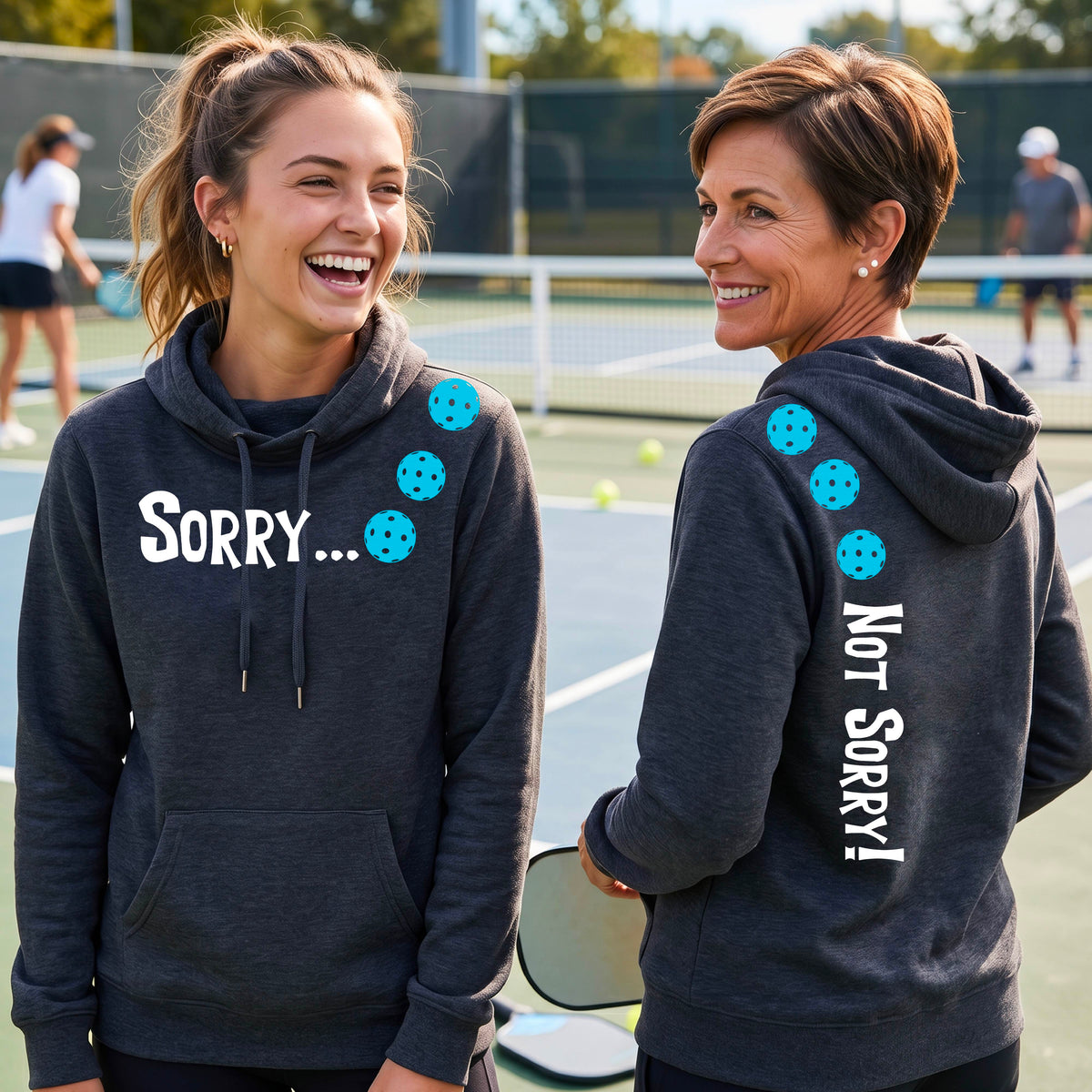 Two women on a pickleball court wearing navy blue pullover hoodies with a front/back graphic design.  The front features the playful word “SORRY…” in bold white lettering, accented with three colorful pickleballs arranged diagonally across the upper chest.  The back design features “NOT SORRY!” in bold white lettering running vertically down the upper back, with three matching colorful pickleballs positioned above lettering.  