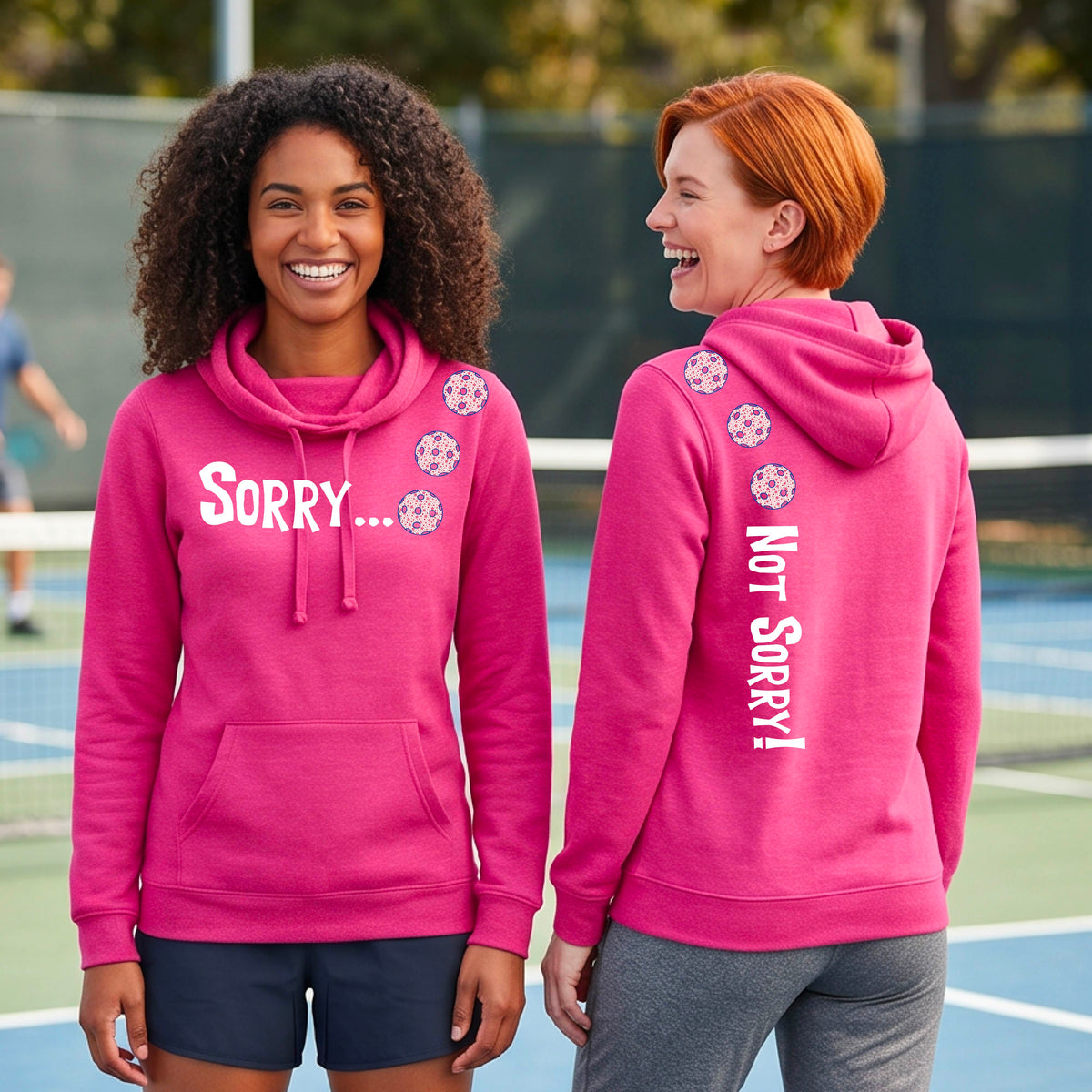 Two women on a pickleball court wearing pink pullover hoodies with a front/back graphic design.  The front features the playful word “SORRY…” in bold white lettering, accented with three colorful pickleballs arranged diagonally across the upper chest.  The back design features “NOT SORRY!” in bold white lettering running vertically down the upper back, with three matching colorful pickleballs positioned above lettering.  