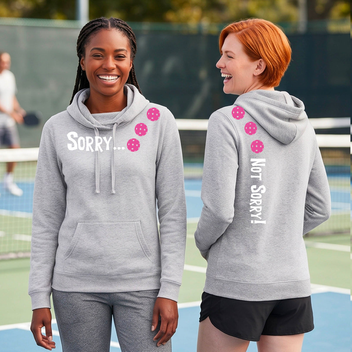 Two women on a pickleball court wearing gray pullover hoodies with a front/back graphic design.  The front features the playful word “SORRY…” in bold white lettering, accented with three colorful pickleballs arranged diagonally across the upper chest.  The back design features “NOT SORRY!” in bold white lettering running vertically down the upper back, with three matching colorful pickleballs positioned above lettering.  