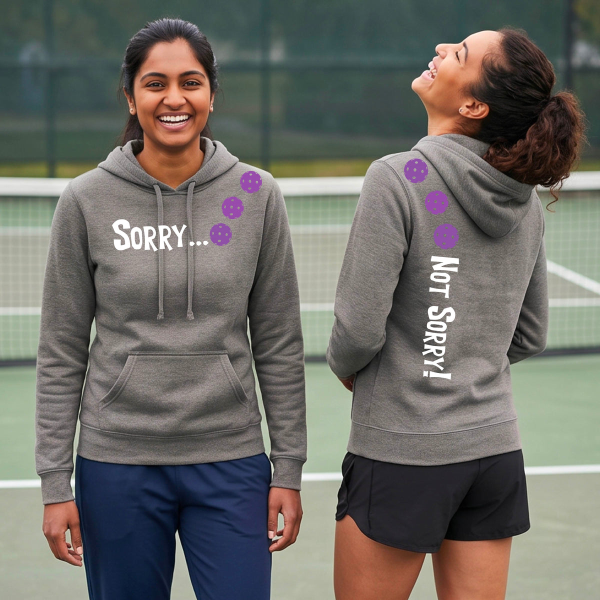 Two women on a pickleball court wearing gray pullover hoodies with a front/back graphic design.  The front features the playful word “SORRY…” in bold white lettering, accented with three colorful pickleballs arranged diagonally across the upper chest.  The back design features “NOT SORRY!” in bold white lettering running vertically down the upper back, with three matching colorful pickleballs positioned above lettering.  