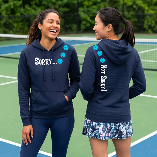 Two women on a pickleball court wearing navy blue pullover hoodies with a front/back graphic design.  The front features the playful word “SORRY…” in bold white lettering, accented with three colorful pickleballs arranged diagonally across the upper chest.  The back design features “NOT SORRY!” in bold white lettering running vertically down the upper back, with three matching colorful pickleballs positioned above lettering.  