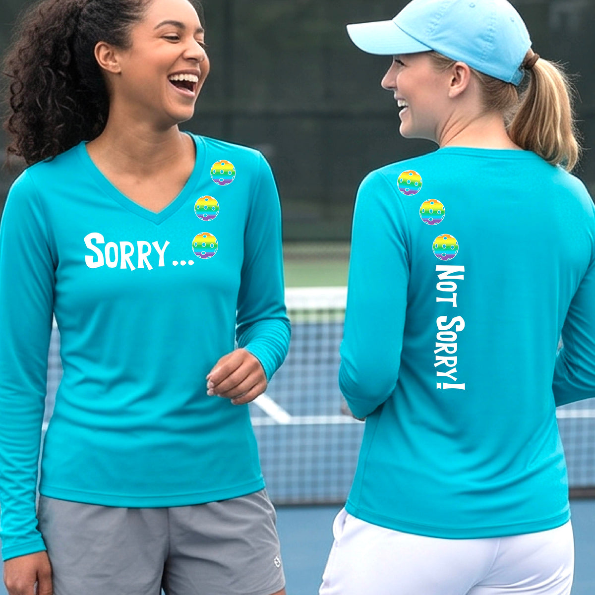 Two women on a pickleball court wearing bright blue long-sleeve v-neck t-shirts with a front/back graphic design.  The front features the playful word “SORRY…” in bold white lettering, accented with three colorful pickleballs arranged diagonally across the upper chest.  The back design features “NOT SORRY!” in bold white lettering running vertically down the upper back, with three matching colorful pickleballs positioned above lettering. 