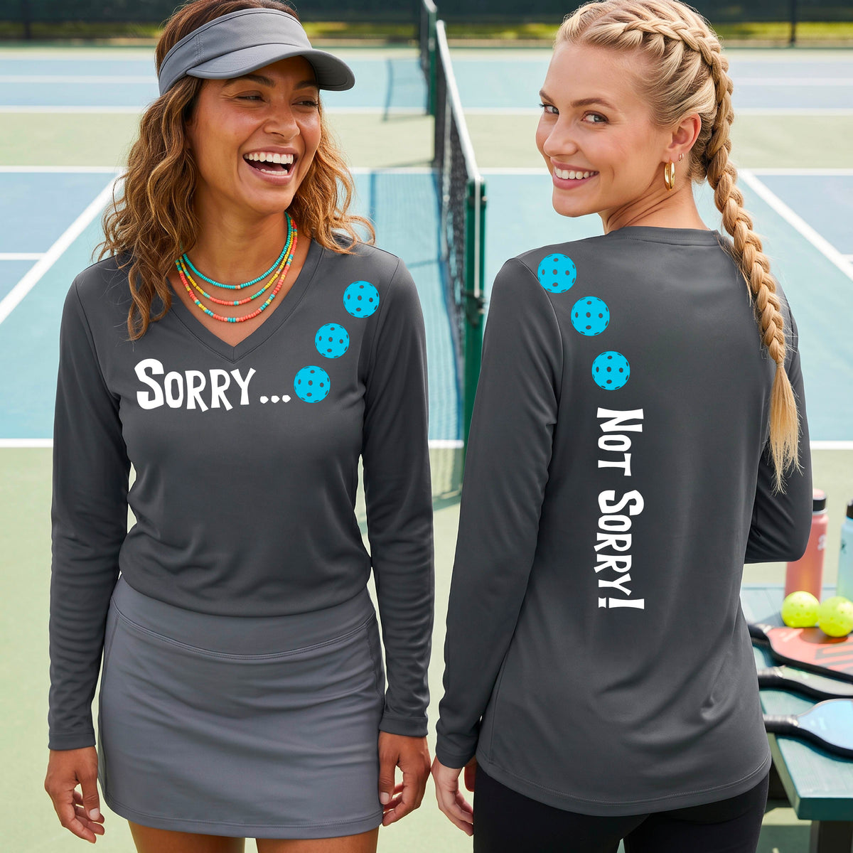 Two women on a pickleball court wearing dark gray long-sleeve v-neck t-shirts with a front/back graphic design.  The front features the playful word “SORRY…” in bold white lettering, accented with three colorful pickleballs arranged diagonally across the upper chest.  The back design features “NOT SORRY!” in bold white lettering running vertically down the upper back, with three matching colorful pickleballs positioned above lettering. 