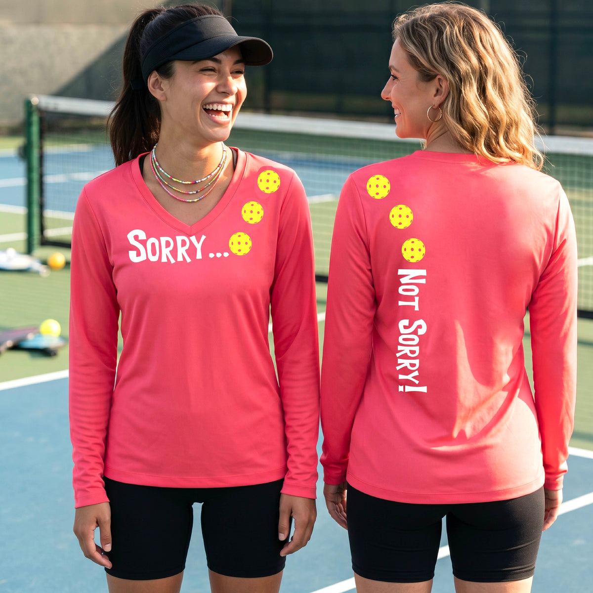 Two women on a pickleball court wearing coral pink long-sleeve v-neck t-shirts with a front/back graphic design.  The front features the playful word “SORRY…” in bold white lettering, accented with three colorful pickleballs arranged diagonally across the upper chest.  The back design features “NOT SORRY!” in bold white lettering running vertically down the upper back, with three matching colorful pickleballs positioned above lettering. 