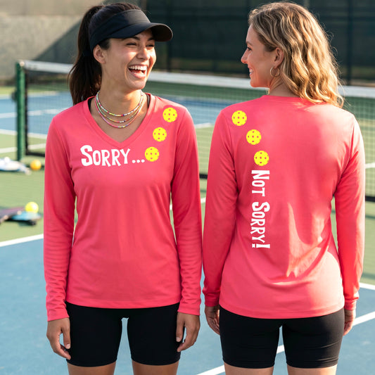 Two women on a pickleball court wearing coral pink long-sleeve v-neck t-shirts with a front/back graphic design.  The front features the playful word “SORRY…” in bold white lettering, accented with three colorful pickleballs arranged diagonally across the upper chest.  The back design features “NOT SORRY!” in bold white lettering running vertically down the upper back, with three matching colorful pickleballs positioned above lettering. 