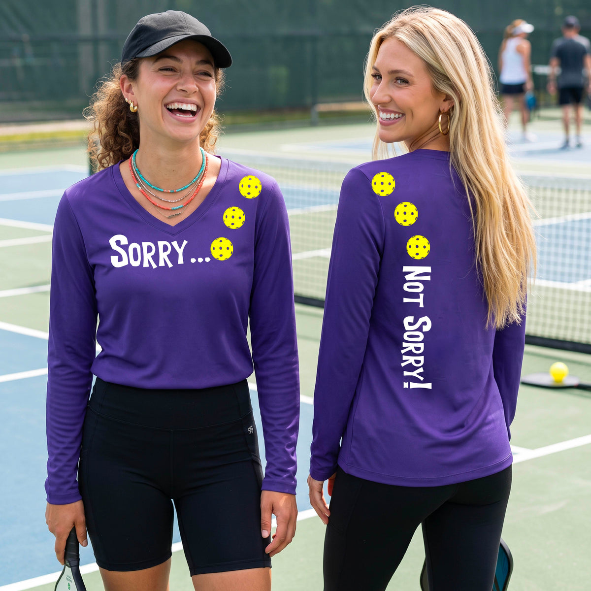 Two women on a pickleball court wearing purple long-sleeve v-neck t-shirts with a front/back graphic design.  The front features the playful word “SORRY…” in bold white lettering, accented with three colorful pickleballs arranged diagonally across the upper chest.  The back design features “NOT SORRY!” in bold white lettering running vertically down the upper back, with three matching colorful pickleballs positioned above lettering. 
