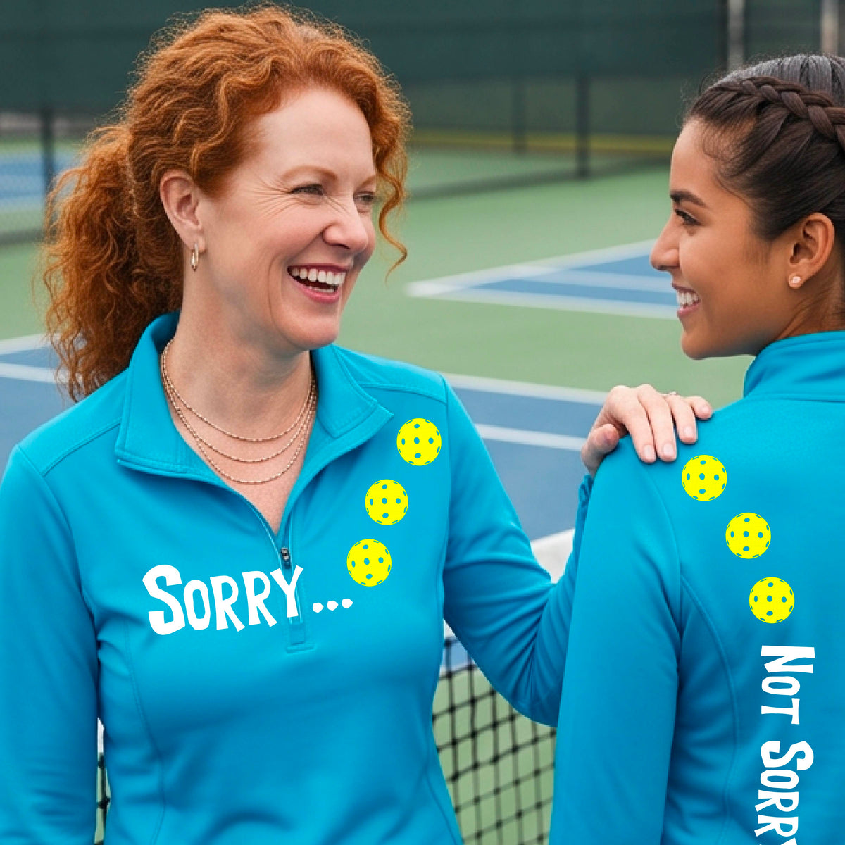 Two women on a pickleball court wearing bright blue quarter-zip shirts with a front/back graphic design.  The front features the playful word “SORRY…” in bold white lettering, accented with three colorful pickleballs arranged diagonally across the upper chest.  The back design features “NOT SORRY!” in bold white lettering running vertically down the upper back, with three matching colorful pickleballs positioned above lettering.