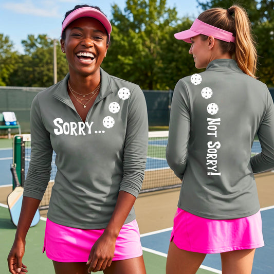 Two women on a pickleball court wearing black quarter-zip shirts with a front/back graphic design.  The front features the playful word “SORRY…” in bold white lettering, accented with three colorful pickleballs arranged diagonally across the upper chest.  The back design features “NOT SORRY!” in bold white lettering running vertically down the upper back, with three matching colorful pickleballs positioned above lettering.  