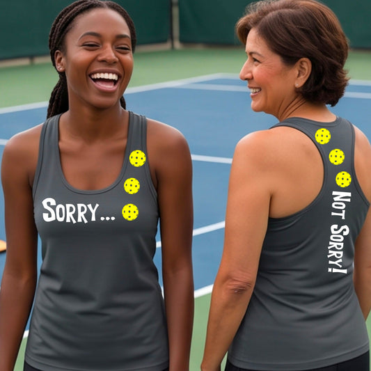 Two women on a pickleball court wearing gray racer-back tank tops with a front/back graphic design.  The front features the playful word “SORRY…” in bold white lettering, accented with three colorful pickleballs arranged diagonally across the upper chest.  The back design features “NOT SORRY!” in bold white lettering running vertically down the upper back, with three matching colorful pickleballs positioned above letters. 
