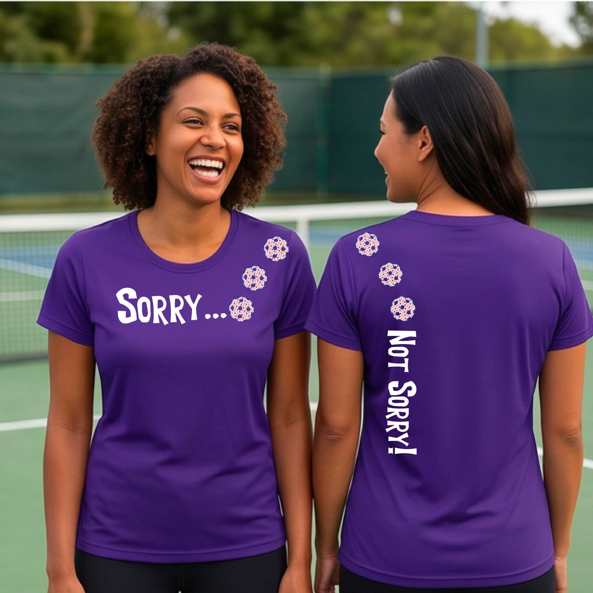 Two women on a pickleball court wearing purple short-sleeve t-shirts with a front/back graphic design.  The front features the playful word “SORRY…” in bold white lettering, accented with three colorful pickleballs arranged diagonally across the upper chest.  The back design features “NOT SORRY!” in bold white lettering running vertically down the upper back, with three matching colorful pickleballs positioned above lettering.  
